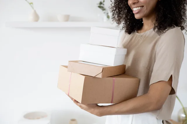 Woman with curly hair carrying stack of packaging boxes in a bright room. Cross-border checkout